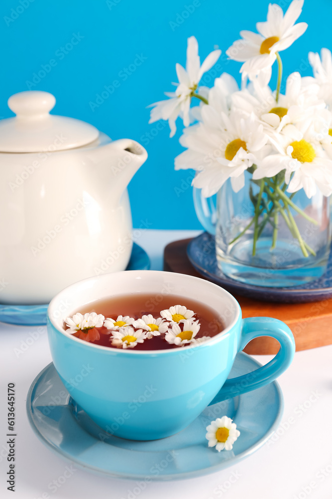 Wooden board with teapot, cup of natural chamomile tea and flowers on white table near blue wall