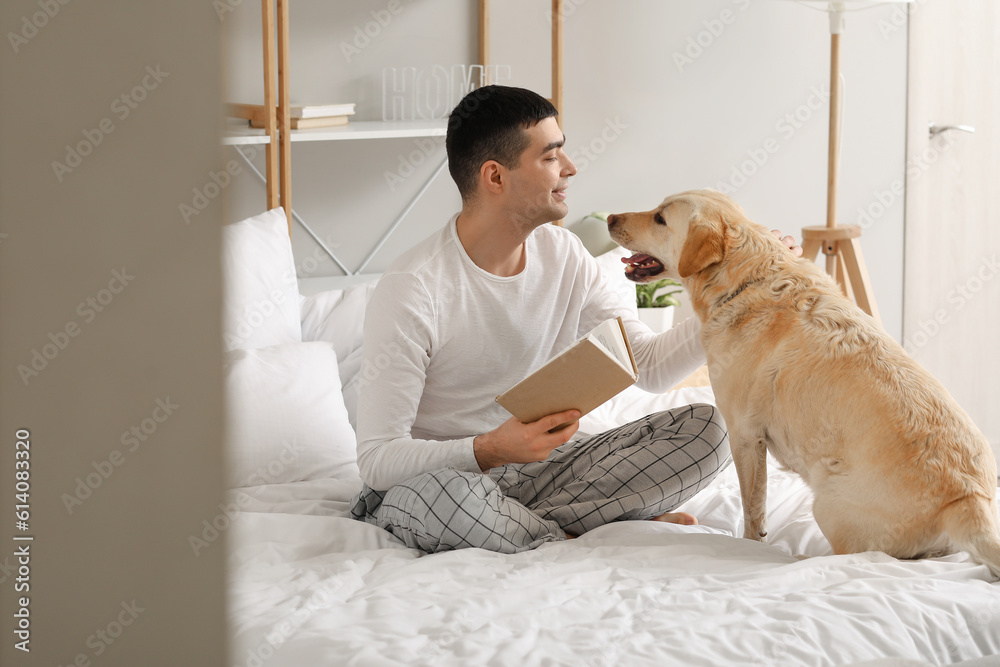 Young man with book and cute Labrador dog sitting in bedroom