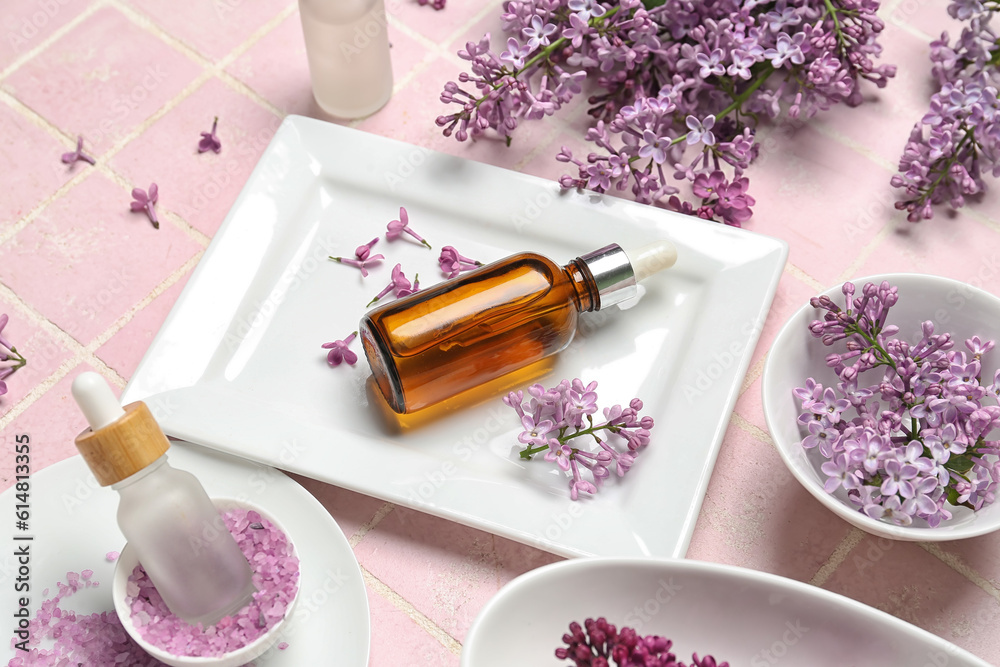 Bottles of lilac essential oil, sea salt and flowers on pink background, closeup