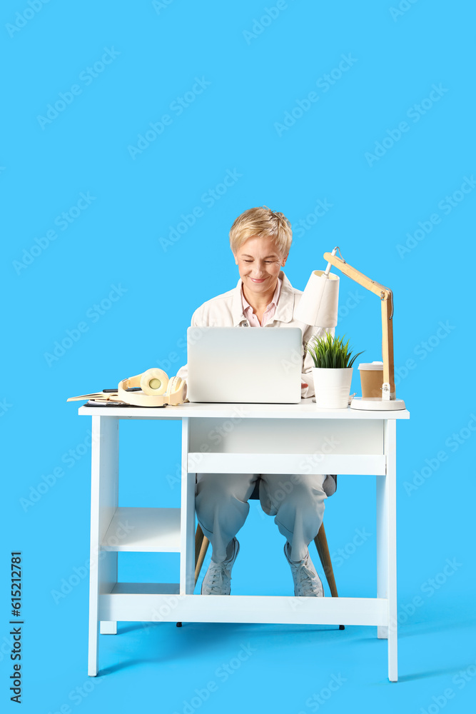 Mature female programmer with laptop at table on blue background