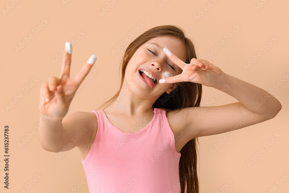 Little girl with sunscreen cream on her face showing victory gesture against beige background