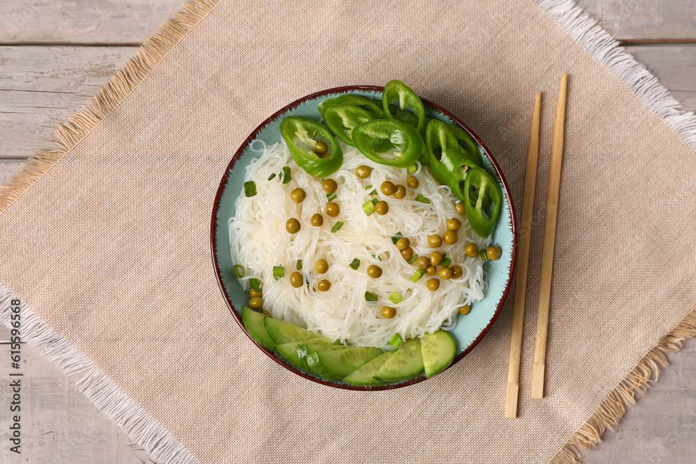 Bowl of tasty rice noodles with chili pepper, green peas and cucumber on light wooden background