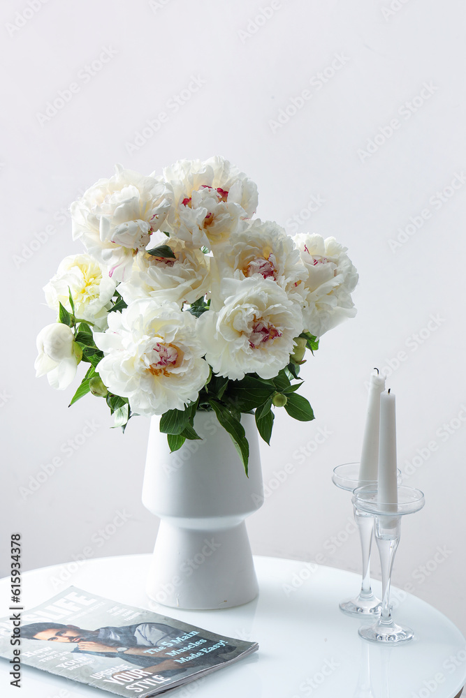 Vase of white peonies with candles and magazine on coffee table near wall