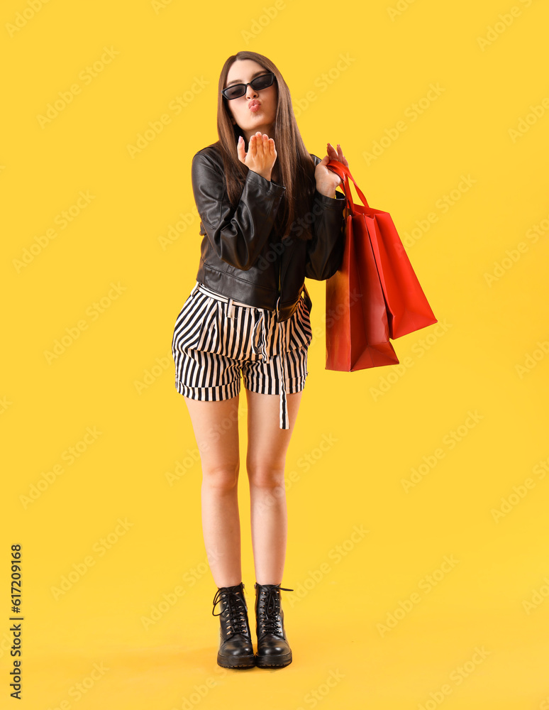 Young woman with shopping bags blowing kiss on yellow background