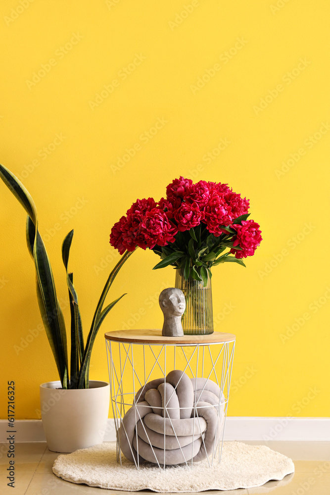 Vase of red peonies with figurine and houseplant near yellow wall