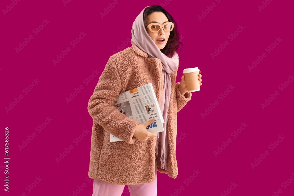 Stylish young woman in teddy coat with cup of coffee and newspaper on pink background
