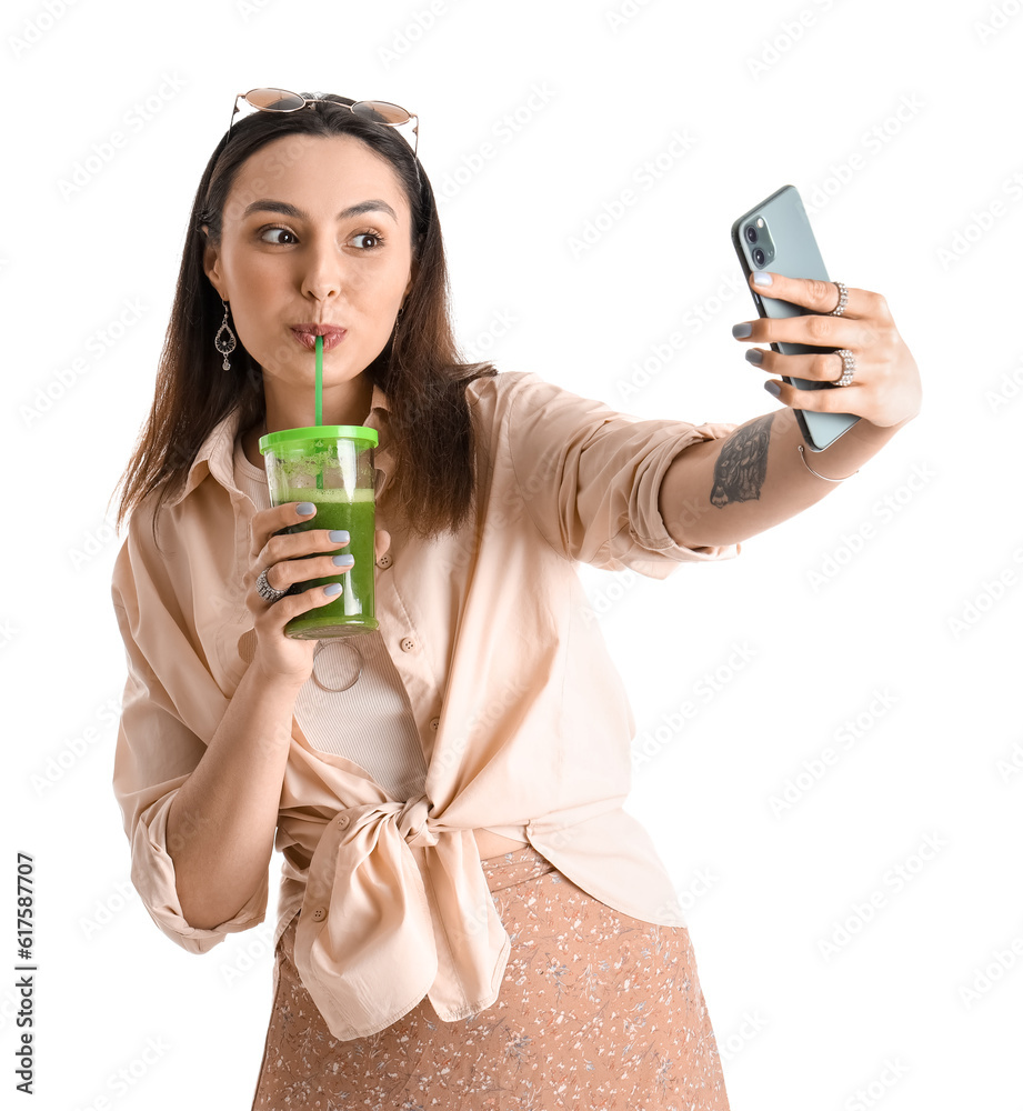 Young woman with glass of vegetable juice taking selfie on white background