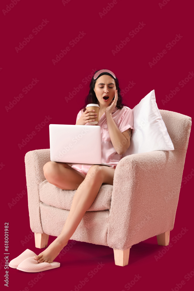 Sleepy young woman with cup of coffee using laptop in armchair on pink background