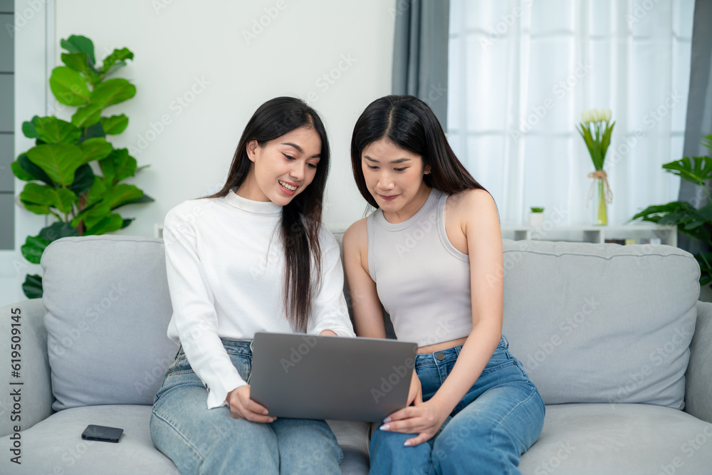 Two young asian women shopping online shopping on laptop while sitting on sofa together in living ro