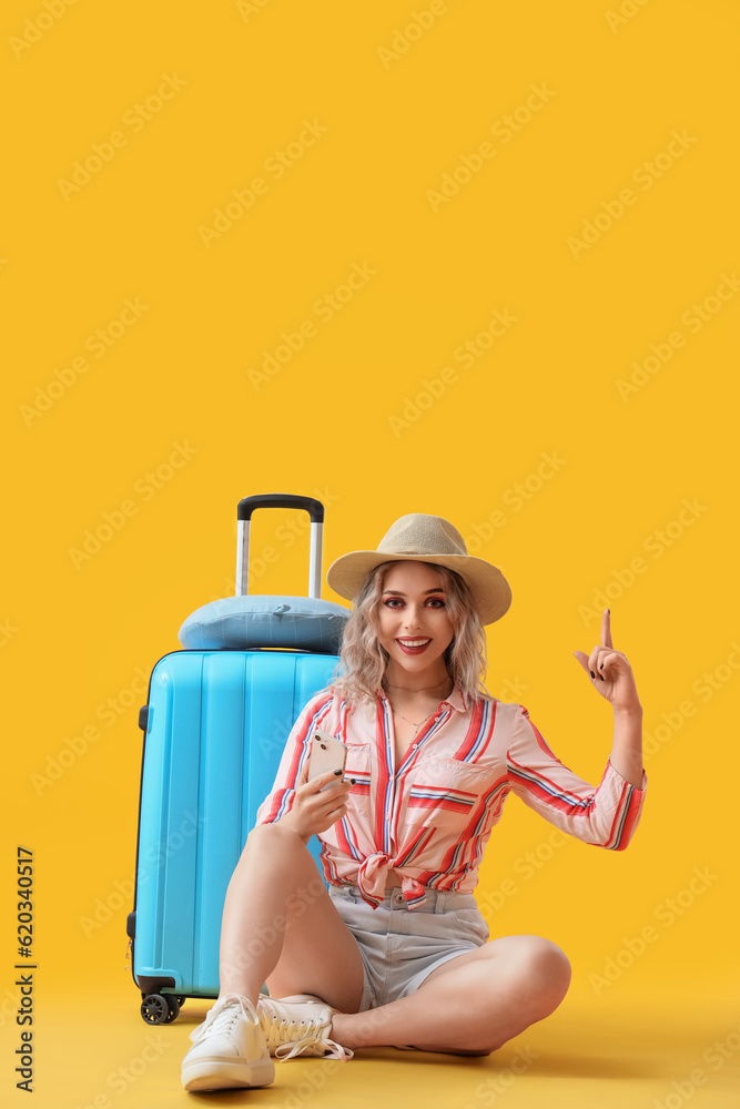Young woman with mobile phone and suitcase pointing at something on yellow background
