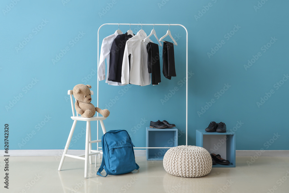 Stylish school uniform hanging on rack, backpack, shoes and pouf near blue wall in room