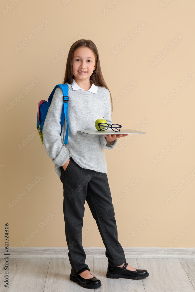 Little schoolgirl with laptop, apple and eyeglasses near beige wall