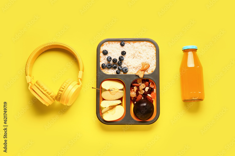 Bottle of juice, headphones and lunchbox with tasty food on yellow background
