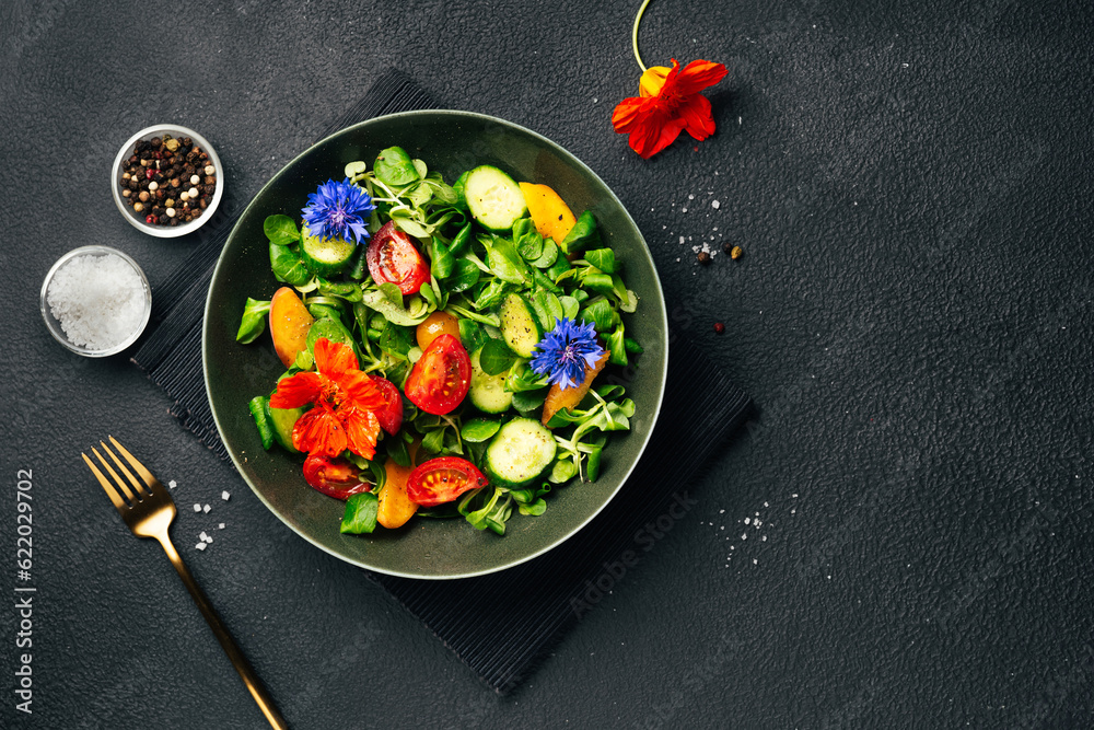 Green salad with fresh cucumbers, tomato, peach and nasturtium flowers on black background. Top view
