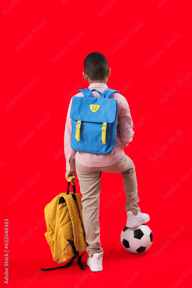 Little African-American schoolboy with soccer ball and backpacks on red background, back view