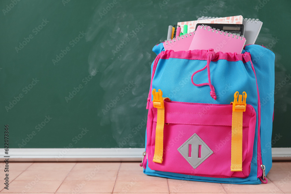 Colorful school backpack with notebooks, calculator and markers on beige tile table near green chalk