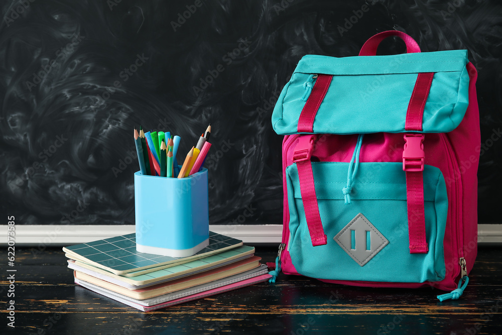 Colorful school backpack with notebooks, cup of pencils and markers on wooden table near black chalk
