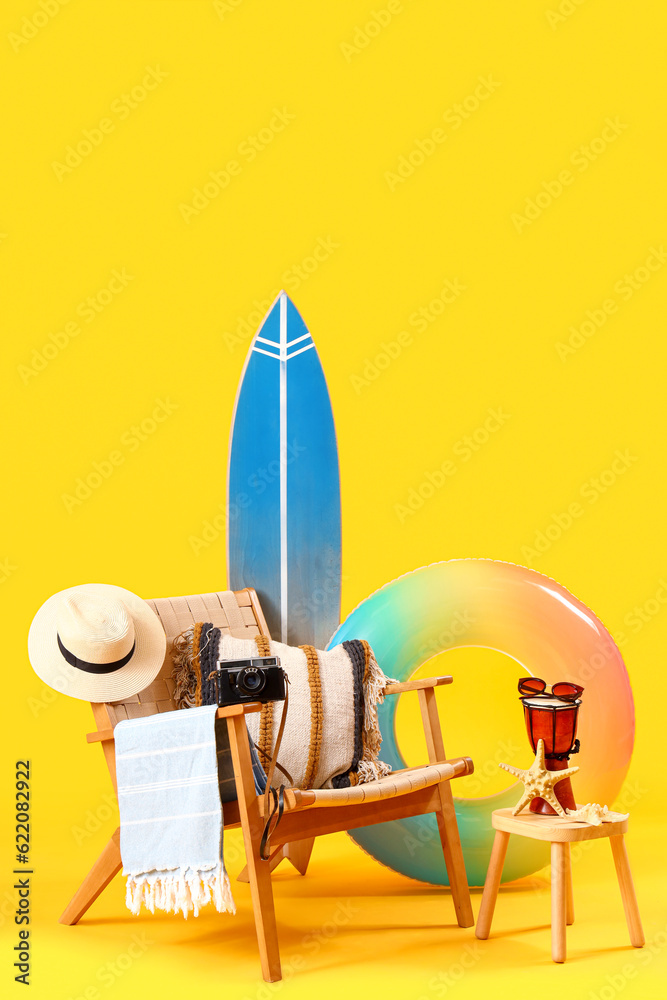 Chair with surfboard, fresh orange juice on stool and beach accessories against yellow background. T