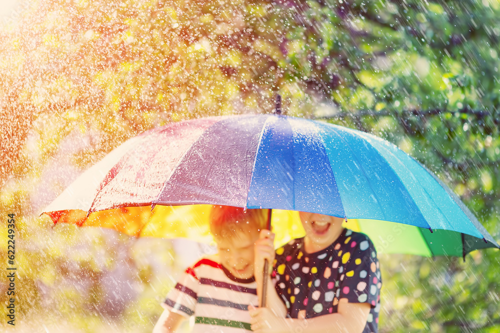 Boy and girl standing outdoors in rainy day under colourful umbrella.