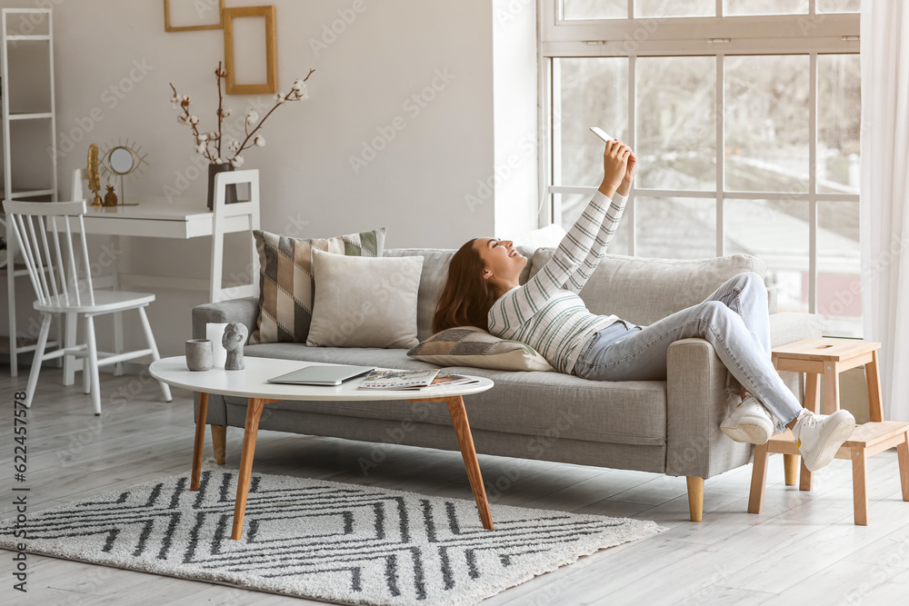 Young woman with mobile phone taking selfie on grey couch in living room
