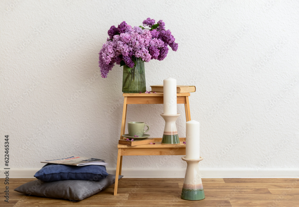Vase with beautiful lilac flowers, cup on step ladder, pillows and candles near light wall in room