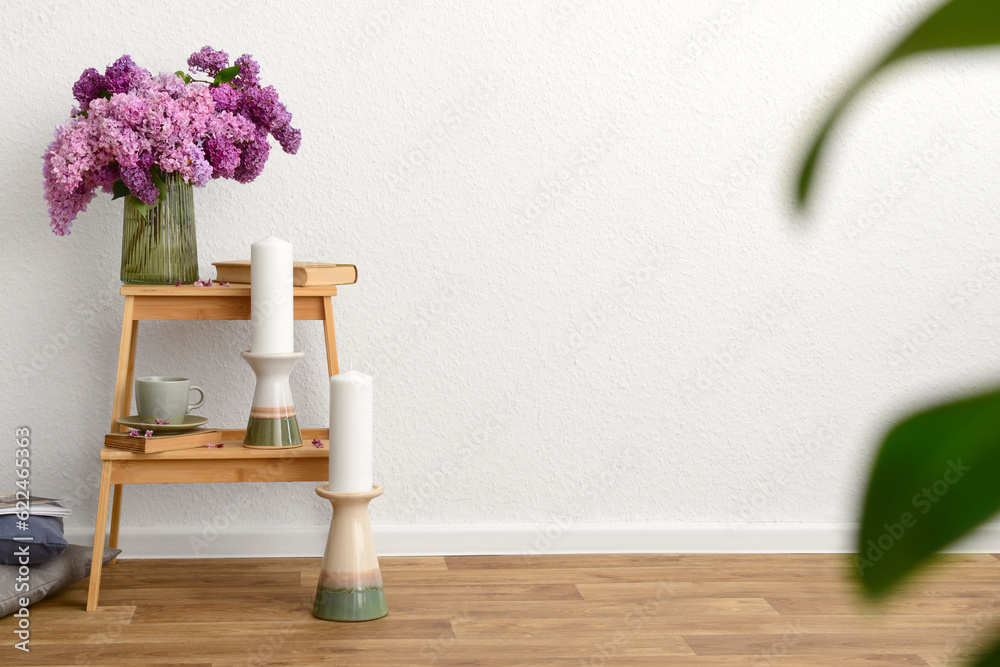 Vase with beautiful lilac flowers, cup and candles on step ladder near light wall in room