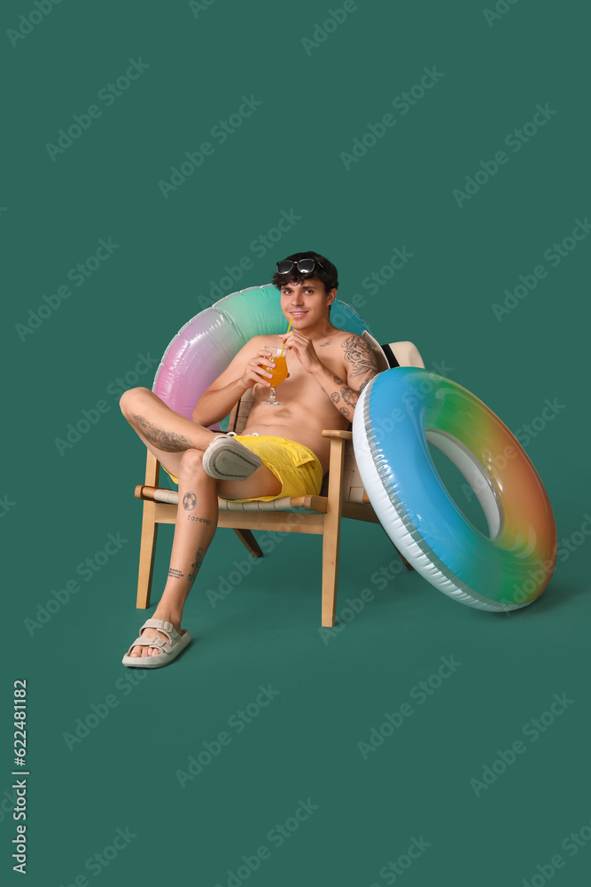 Young man drinking summer cocktail in chair and swim rings on green background