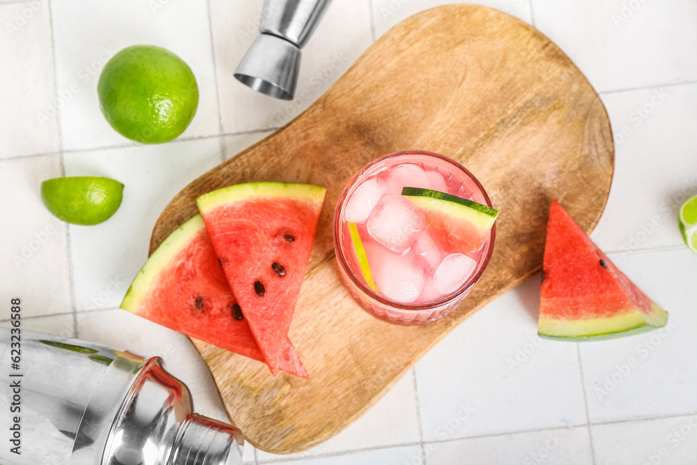 Board with glass of tasty watermelon cocktail, shaker, jigger and lime on white tile background
