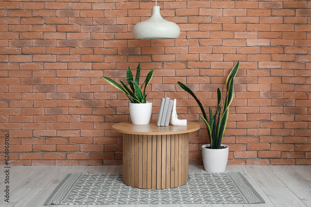 Wooden coffee table with houseplants and stylish holder for books in room near brick wall