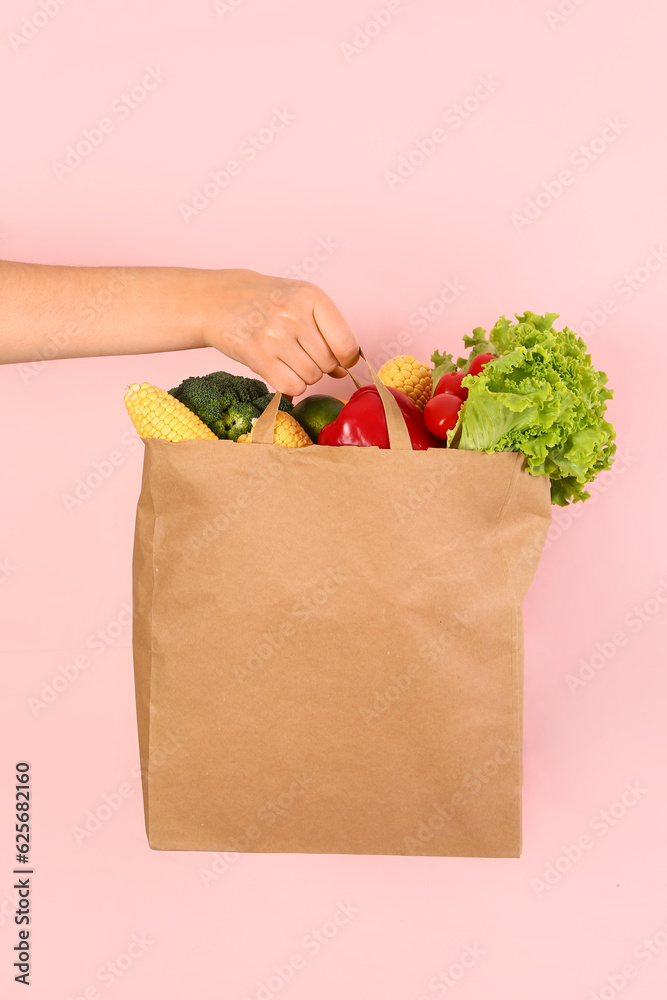 Female hand with paper bag full of fresh vegetables on pink background