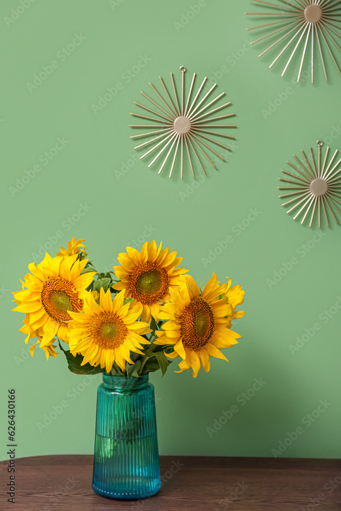 Vase with beautiful sunflowers on table and decor hanging on green wall in room