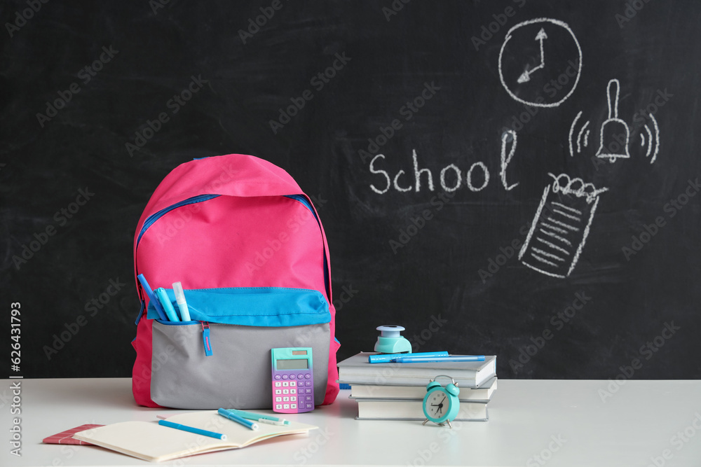 Colorful school backpack with different stationery on white table near black chalkboard