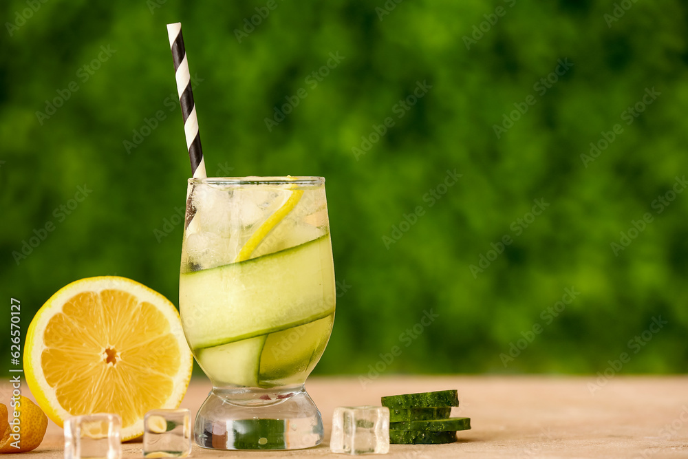 Glass of lemonade with cucumber and ice cubes on beige table outdoors