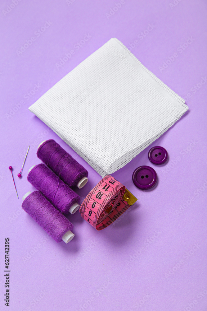 Canvas with buttons, thread spools and measuring tape on lilac background
