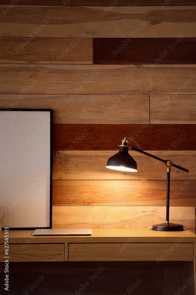 Desk with glowing lamp, modern laptop and blank frame near wooden wall