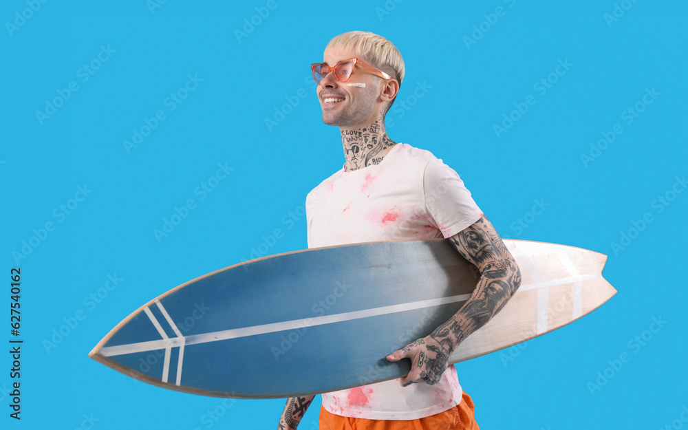 Young tattooed man with surfboard on light blue background