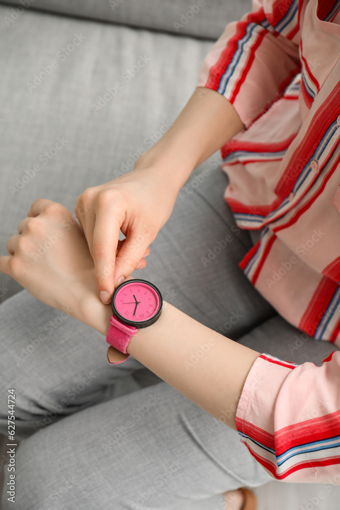 Young woman sitting on sofa and looking at stylish wristwatch in room, closeup