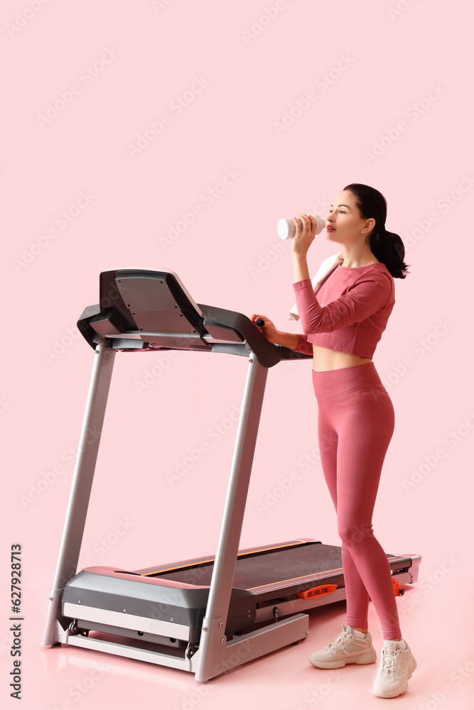 Beautiful woman with treadmill drinking water on pink background