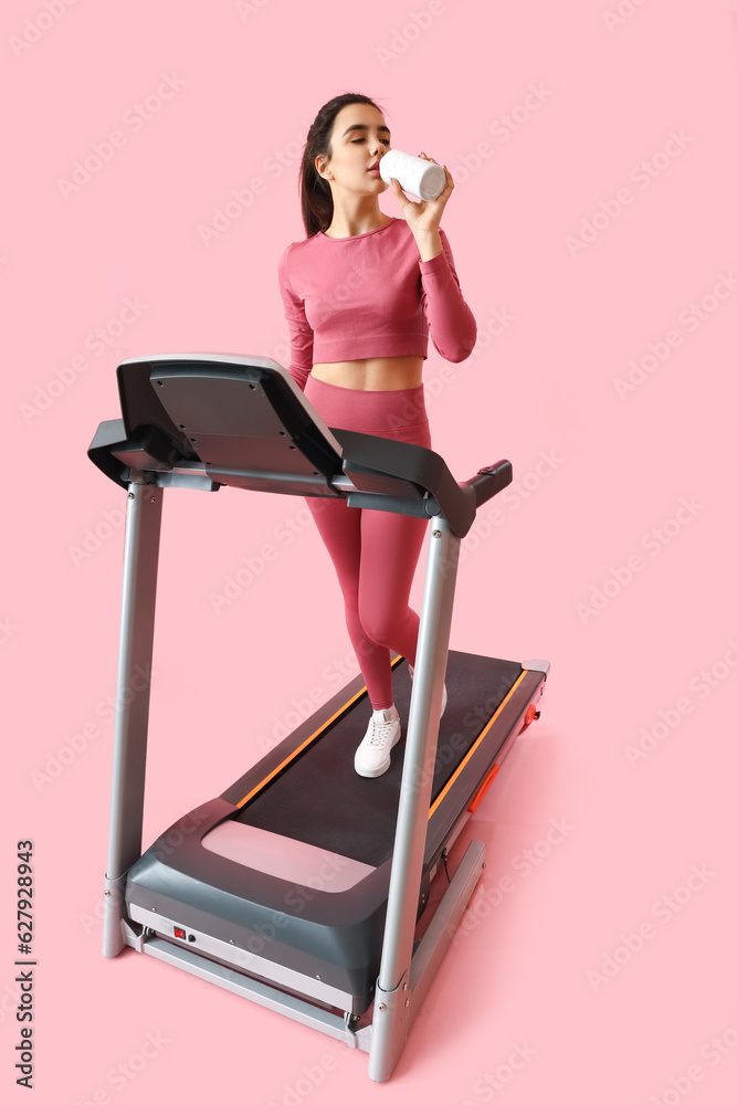 Sporty young woman drinking water on treadmill against pink background