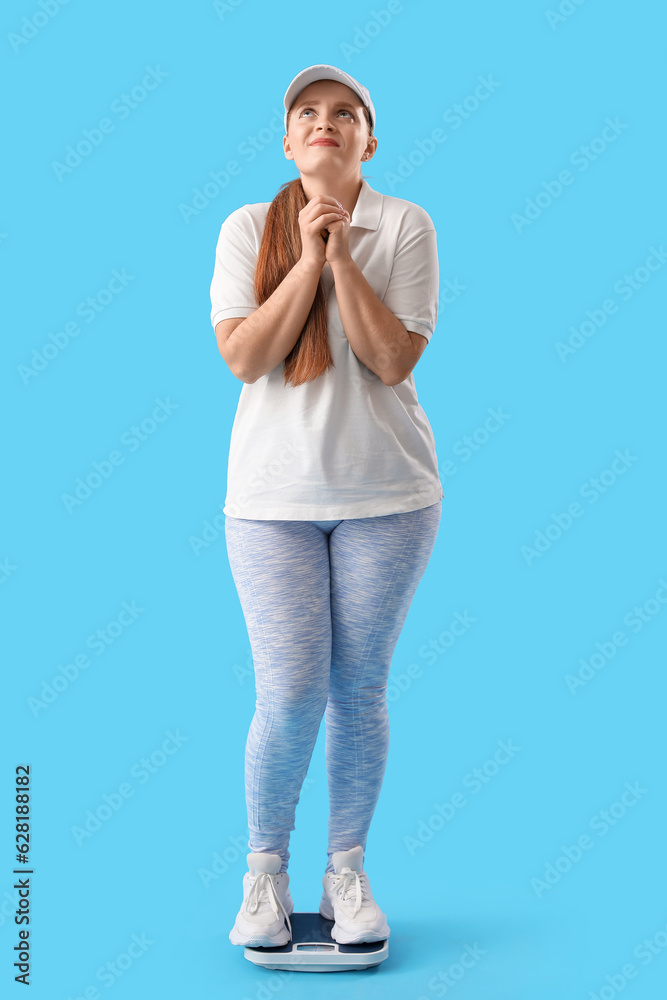 Worried young woman measuring her weight on scales against blue background