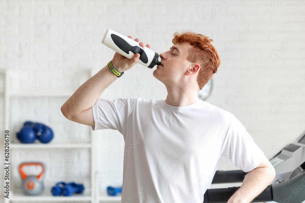 Young redhead man drinking water after training in gym