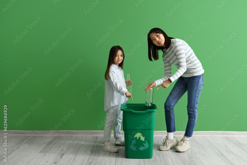 Asian mother with her little daughter throwing glass bottles in recycle bin near green wall
