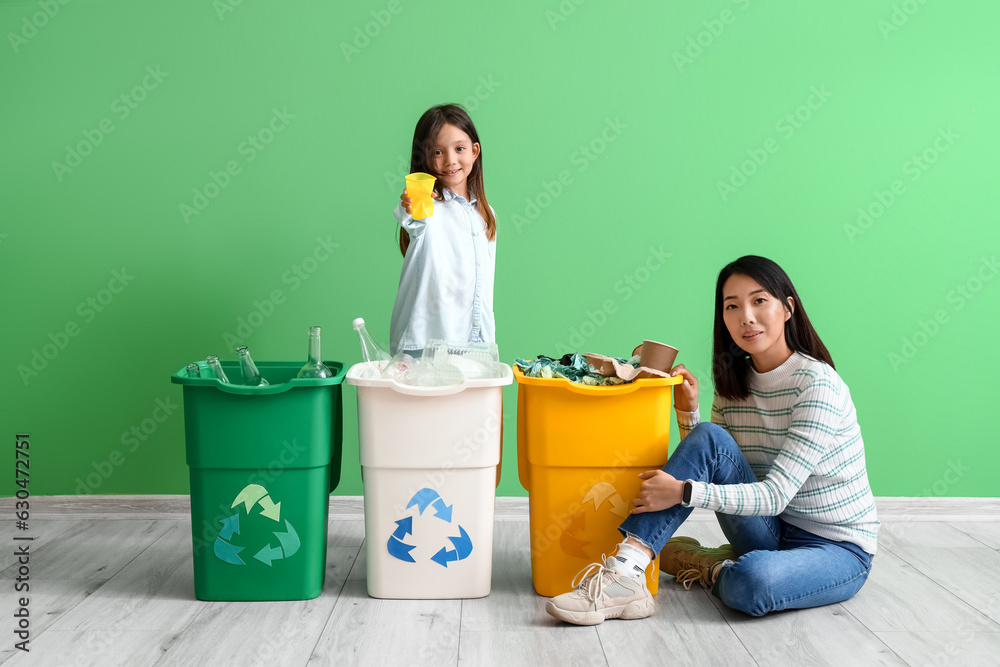 Asian mother with her little daughter sorting garbage in recycle bins near green wall