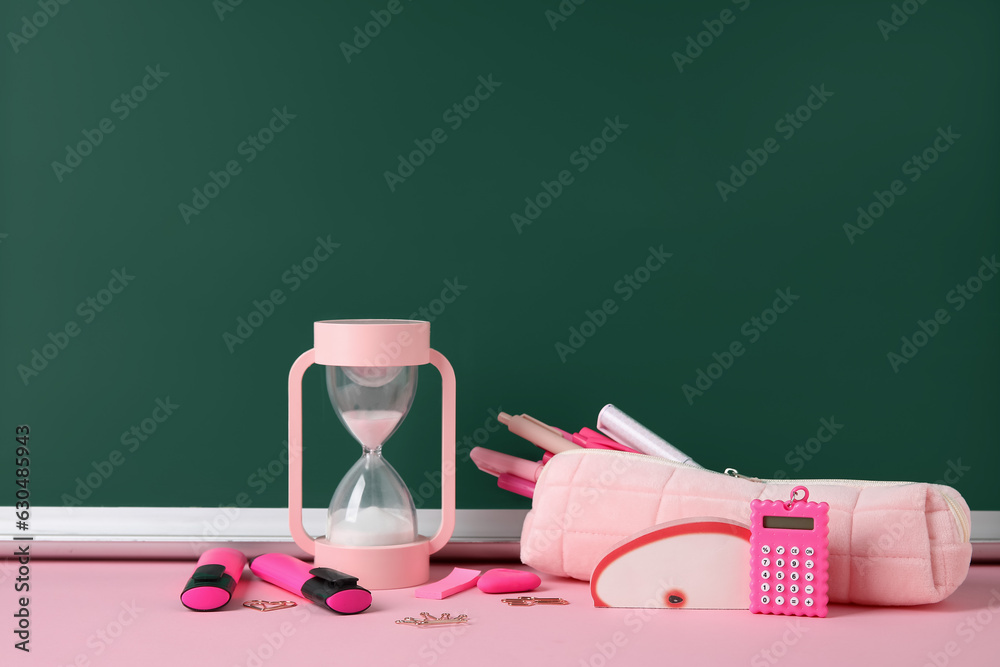 Pencil case with different school stationery and hourglass on pink table near blackboard