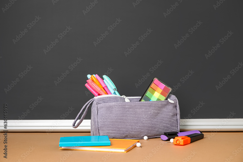 Pencil case with different school stationery on table near blackboard