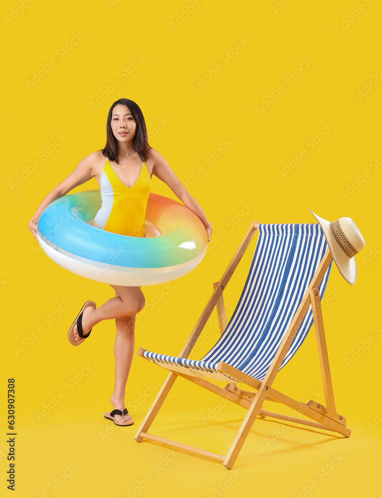 Beautiful Asian woman with swim ring and deck chair on yellow background