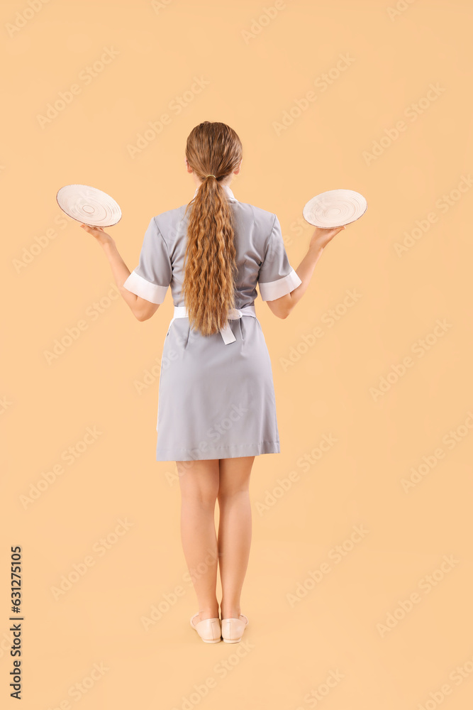 Beautiful young waitress with empty plates on beige background, back view