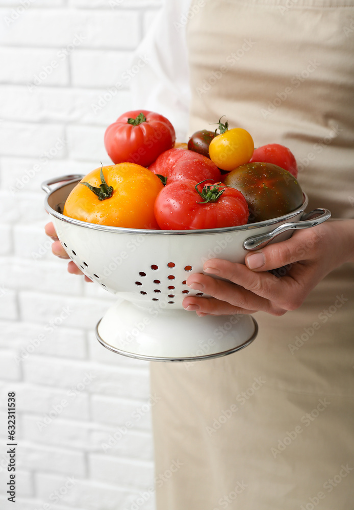 Woman holding colander with different fresh tomatoes on white brick background, closeup