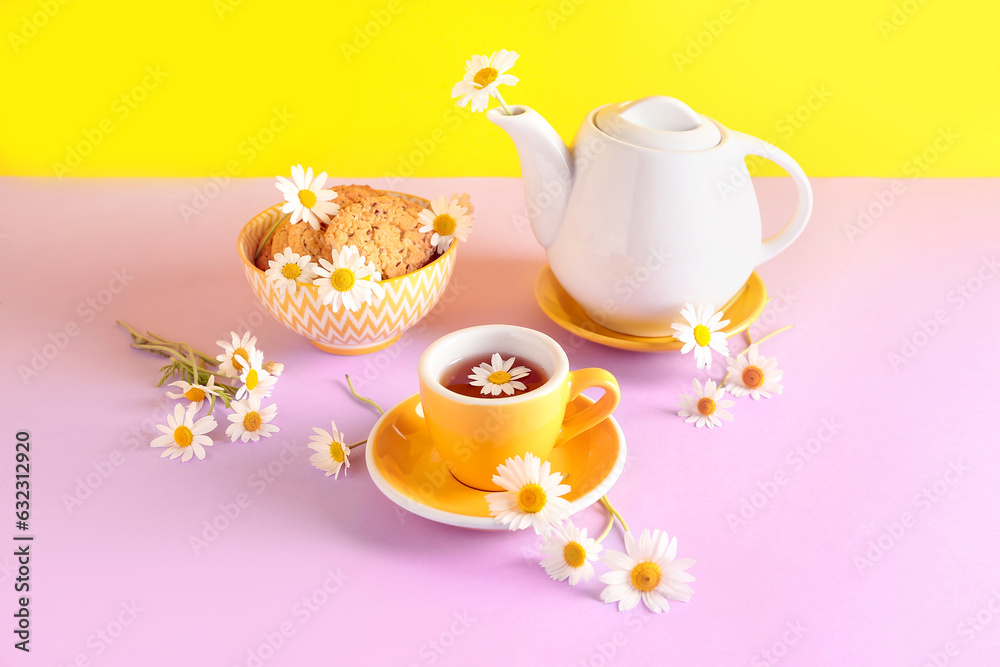 Teapot with cup of natural chamomile tea, cookies and flowers on pink table near yellow wall