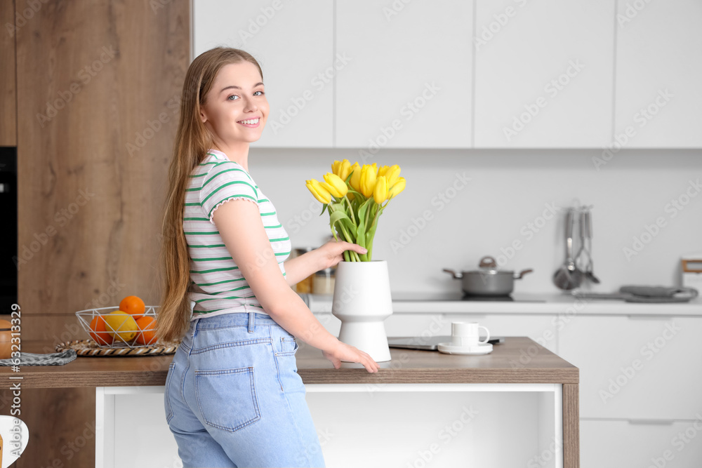 Beautiful young woman putting bouquet of yellow tulip flowers into vase in modern kitchen